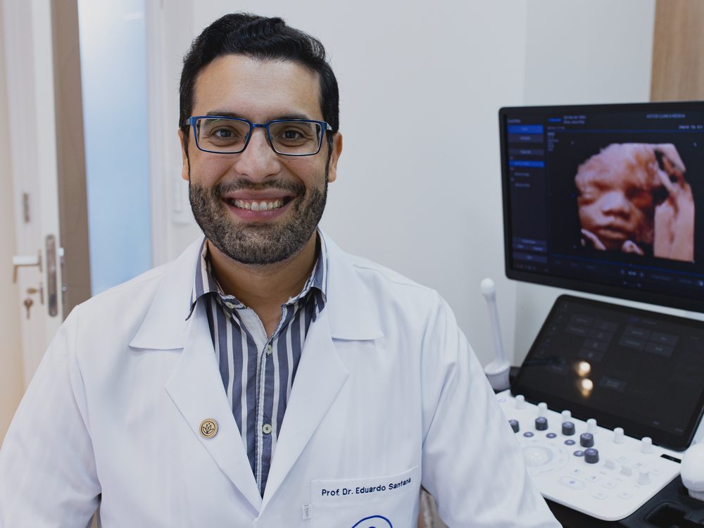 Dr. Eduardo Santana, obstetra, de jaleco, sorrindo ao lado do aparelho de ultrassom